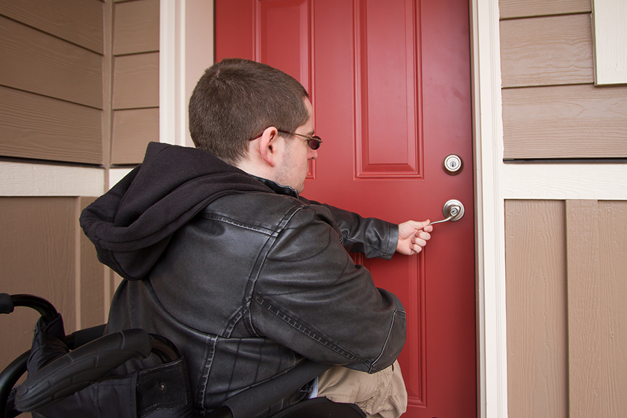 A man in a black jacket using a wheelchair uses a silver-colored lever-style door handle to open the red door into a home.